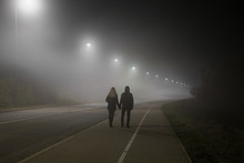 Young Couple Holding Each Other Hands. Slowly Walking Under White Street Lights In Night. Dark Time. Peaceful Atmosphere In Mist. Foggy Air.