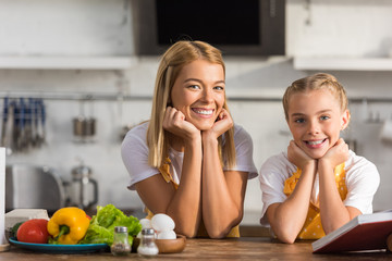 Wall Mural - happy mother and daughter in aprons leaning at kitchen table and smiling at camera