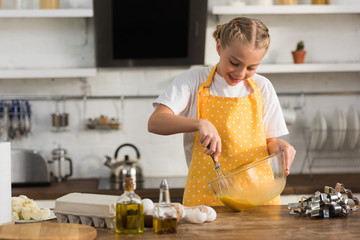 Wall Mural - adorable smiling chuild in apron whisking eggs while cooking in kitchen