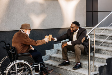 senior disabled man in wheelchair and african american man drinking coffee together and clinking paper cups on street