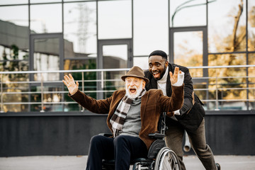 happy senior disabled man in wheelchair and african american man having fun while riding by street