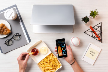 Poster - cropped shot of person eating french fries with ketchup and using smartphone with business charts at workplace