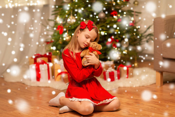 christmas, holidays and childhood concept - girl in red dress hugging teddy bear at home