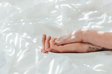 partial view of loving couple holding hands while lying on white bed sheet