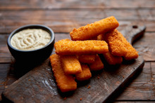 Pile Of Golden Fried Fish Fingers With White Garlic Sauce Placed On Chopping Board On Dark Wooden Background.