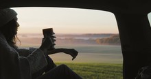 Young woman admiring the sunrise in trunk of car. Girl dressed in woolen socks drinking hot coffee against backdrop of forest panorama in fog . View from inside 4k video