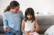 © fizkes - Little adorable daughter and young beautiful mother sitting on couch at home small kid receive from loving smiling mother pink gift box at birthday. Life events and celebration congratulation concept