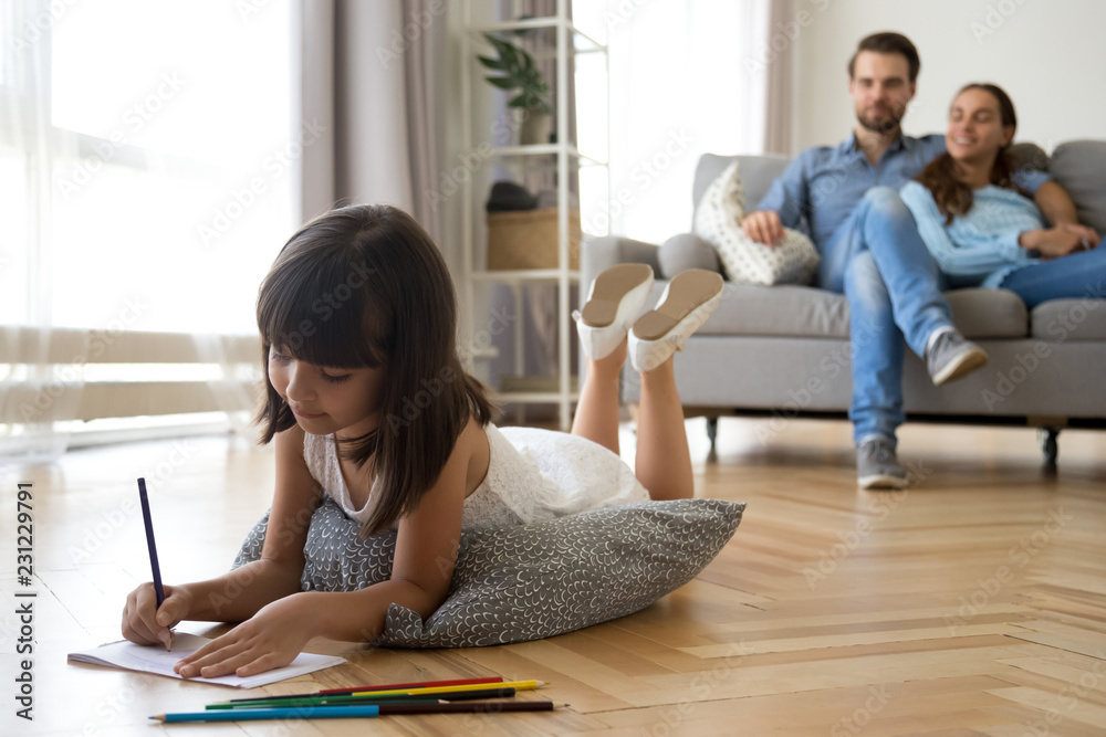 Diverse family in living room at home. Focus on little preschool adorable concentrated daughter lying at soft cushion on warm wooden floor young parents married couple sitting on sofa looking at kid - obrazy, fototapety, plakaty 