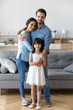 Full length diverse multi-ethnic family married couple wife husband little daughter embracing standing together in living room smiling looking at camera at new modern home feels happy and satisfied.