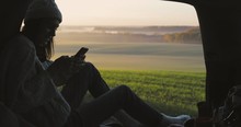 Young woman admiring the sunrise in trunk of car. Girl dressed in woolen clothes reading and texting massages on mobile phone against backdrop of forest panorama in fog . View from inside 4k video