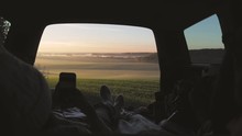 Woman admiring the sunrise in trunk of car. Girl dressed in woolen clothes reading and texting massages on mobile phone against backdrop of forest panorama in fog . View from inside slow motion video