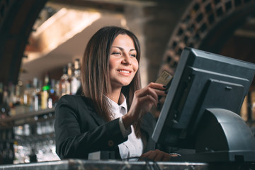 small business, people and service concept - happy woman or waiter or manager in apron at counter with cashbox working at bar or coffee shop