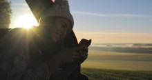 Young woman admiring the sunrise in trunk of car van. Girl dressed in woolen socks taking photo on mobile phone against backdrop of forest panorama in fog . View from inside 4k video