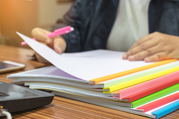 Teacher hand is holding pen for checking student homework assignments on desk in school. Unfinished paperwork stacked in archive with color plastic slide binder bars. Education and business concept.