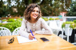 © F8  \ Suport Ukraine - Latin curly female student writing in copybook at sidewalk cafe. Portrait of Caucasian woman wearing white T-shirt sitting at table by university cafe