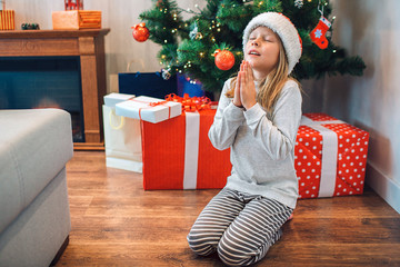 Small girl sitting on her knees and praying. She keeps eyes closed and holds hands togther. Child wishing and saying thanks. There are boxes wth gifts and Christmas tree behind her.