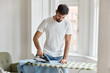 Hard working bearded man dressed in white t shirt, irons shirt on ironing board, takes care of clothes, does household duties, poses against window background. Housework and masculinity concept