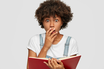 Wall Mural - Surprised frightened black girl covers mouth from amazement, doesnt believe in shocking news, holds textbook, keeps eyes popped out, poses against white background. Facial expressions concept