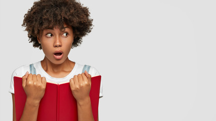 Wall Mural - Shot of shocked schoolgirl has curly hair, openes mouth from surprise, holds textbook, realizes day of exam is coming, poses against white background with free space for your advertisement, promotion