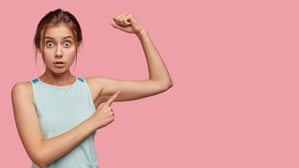 Wall Mural - Horizontal studio shot of surprised European girl indicates at her muscle, stares at camera with eyes popped out, cant believe in such quick results, has outdoor trainings on biceps, likes sport