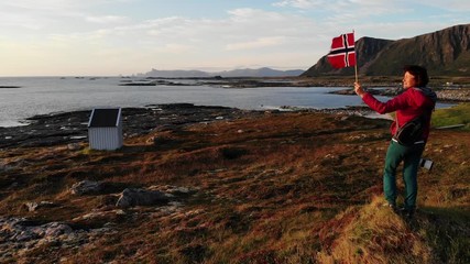 Poster - Aerial view. Tourist woman with waving norwegian flag enjoying sea coast view on rest stop area on Andoya island near Nordmela village Vesteralen archipelago, Norway