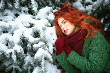 Wall Mural - Close up portrait of a beautiful red-haired girl wearing green coat, vinous snood scarf and mittens leaning on snowy fir tree bough