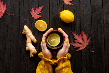 Mug of tea in female hands. Lemon, ginger and autumn leaves. Cold treatment. Wooden background. Top view