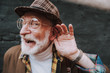What. Close up portrait of stylish pensioner with playful glance holding palm near his ear and smiling