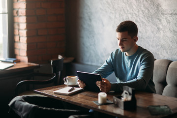 guy is sitting in a cafe and using a tablet. student spend time in cafe and drink coffee. he study a