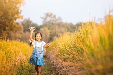 Wall Mural - Cute asian child girl running and playing with toy wooden airplane in the field at sunset time with fun