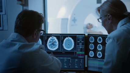 Wall Mural - In Control Room Doctor and Radiologist Discuss Diagnosis while Watching Procedure and Monitors Showing Brain Scans Results, In the Background Patient Undergoes MRI or CT Scan Procedure.