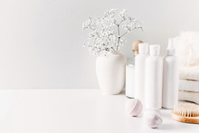 Soft Light Bathroom Decor For Advertising, Design, Cover, Set Of Cosmetic Bottles, Bath Accessories, White Small Flowers In Vase, Towel On White Wooden Shelf. Mock Up,copy Space 