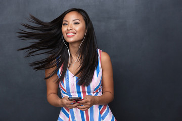 Wall Mural - Image of joyful asian woman 30s wearing earphones listening to music on black smartphone, standing isolated over gray background