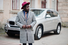Stylish African American Man Model In Gray Coat, Jacket Tie And Red Hat Against Silver Business Suv Car.