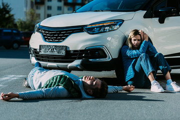 Wall Mural - scared young woman sitting near car and looking at injured man lying on road after traffic collision