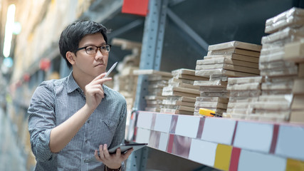 Wall Mural - Young Asian man worker doing stocktaking of product in cardboard box on shelves in warehouse by using digital tablet and pen. Physical inventory count concept