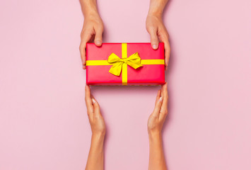 Top view of male and female hands holding red gift box with golden ribbon on pink background Flat lay. Present for birthday, valentine day, Christmas, New Year. Congratulations background copy space.