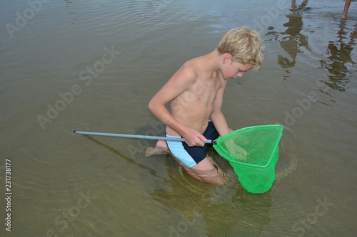 Fotografie, Obraz Blonder Junge sitzt im seichten Wasser am Strand mit einem Fischernetz - Käscher