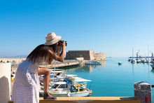 Elegant Young Tourist Visitor Woman Walking On A Sightseeing Tour At Heraklion Venetian Port, Crete, Greece. Venetian Fort At Background.