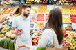 © pressmaster - Confused misunderstanding young male grocer in apron holding container of strawberries shrugging shoulders while talking to customer in farmers market