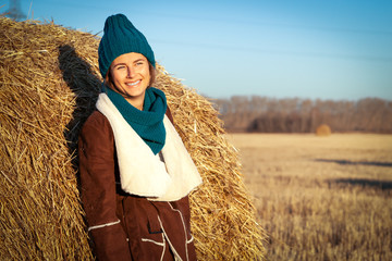 Wall Mural - Young attractive lady in blue knitting hat and brown coat stay under haystack. Smiling beautiful hipster happy woman on field, wearing stylish clothes. Autumn trend, urban stydent style.