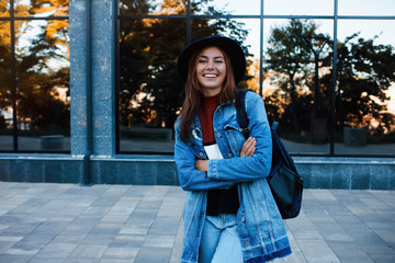 Wall Mural - Hipster girl wearing casual sweater, jeans and backpack posing against glass street wall, minimalist urban clothing style.Stylish happy young woman wearing boyfriend jeans and long jeans coat.