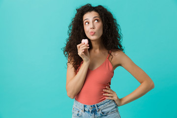 Poster - Photo of gorgeous woman 20s with curly hair smiling and eating macaron biscuit, isolated over blue background