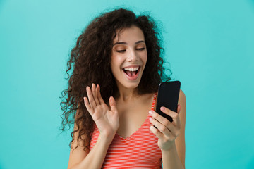 Wall Mural - Photo of joyful woman 20s with curly hair smiling and looking at mobile phone, isolated over blue background