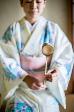 Japanese Woman Wearing Traditional White Kimono With Blue Floral Pattern Kneeling On Floor During Tea Ceremony, Holding A Hishaku, A Bamboo Ladle.