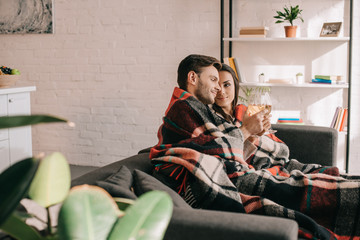 Wall Mural - happy young couple with glasses of white wine relaxing on couch under plaid