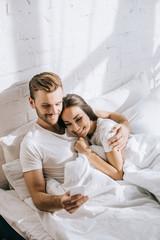 Wall Mural - high angle view of beautiful young couple relaxing in bed in morning and using smartphone