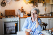 © Marina April - Senior woman is talking on mobile phone at cozy home kitchen. Grandmother has tea or coffee break in cooking. Retired person is using device. Concept of people cyber privacy, connection, communication