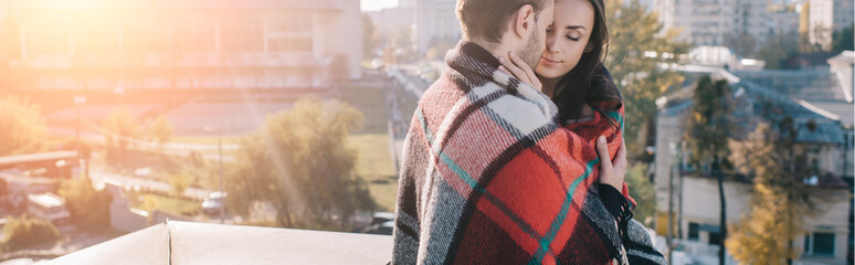 Wall Mural - panoramic shot of beautiful young couple covering in plaid and cuddling on rooftop