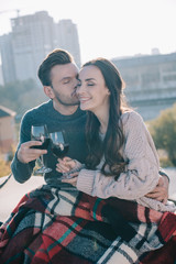 Wall Mural - beautiful young couple covering in plaid and clinking glasses of red wine on rooftop